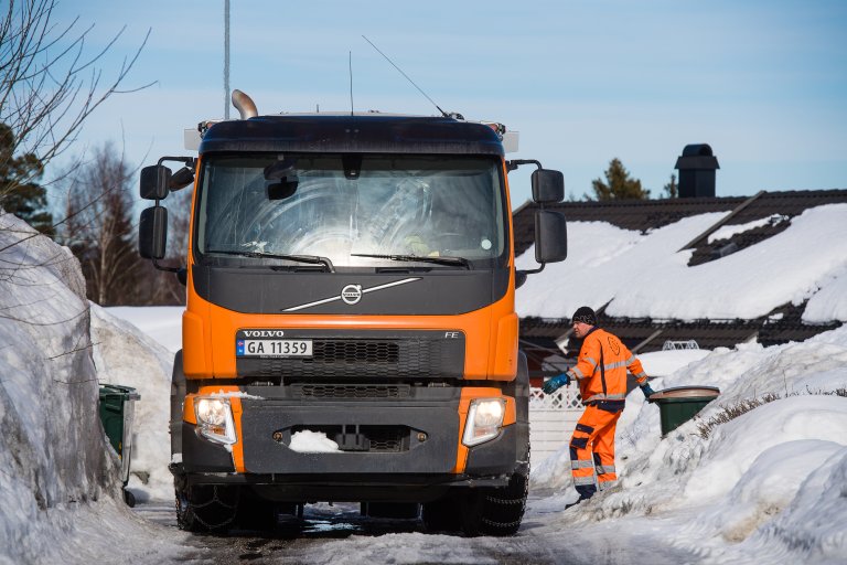 En renovasjonsbil står i en gate med snø på begge sider, mens en renovatør triller frem en beholder.