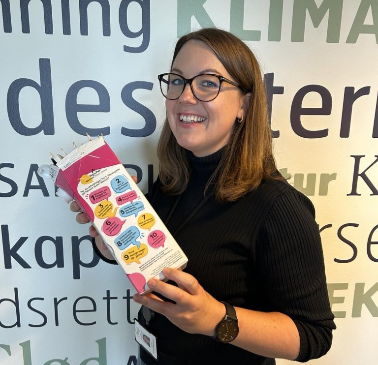 En kvinne holder opp en melkekartong og smiler.