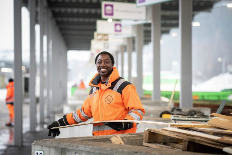 En mann i oransje arbeidsklær med RfD-logo på smiler foran avfallskonteineren på Mile gjenvinningsstasjon.