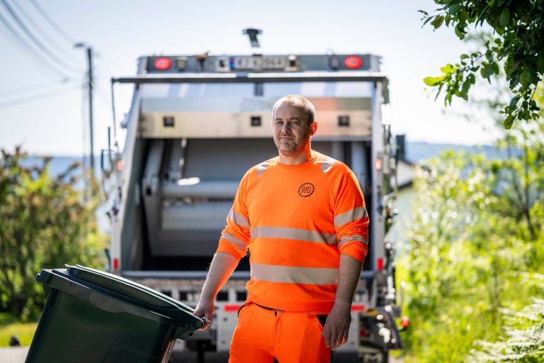 En renovatør i oransje arbeidsklær med RfD-logo på brystet står foran en renovasjonsbil med en avfallsbeholder. Han smiler.